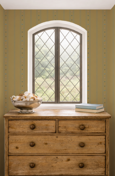 Wooden dresser with a bowl of seashells and books in front of an arched window with diamond-patterned glass on a green wall.