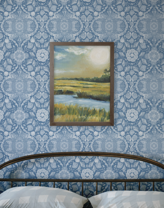 Framed landscape painting on a blue patterned wall above a bed.