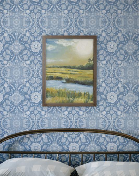 Framed landscape painting on a blue patterned wall above a bed.