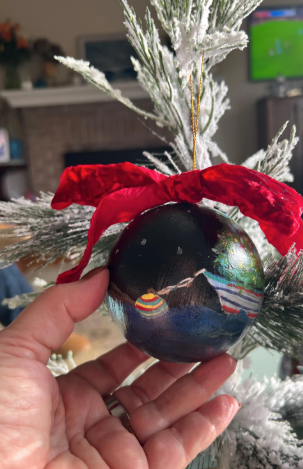Decorative ornament with a red bow held by a hand in front of a Christmas tree.