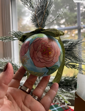 Hand holding a decorative ornament with floral design in front of a window with greenery.