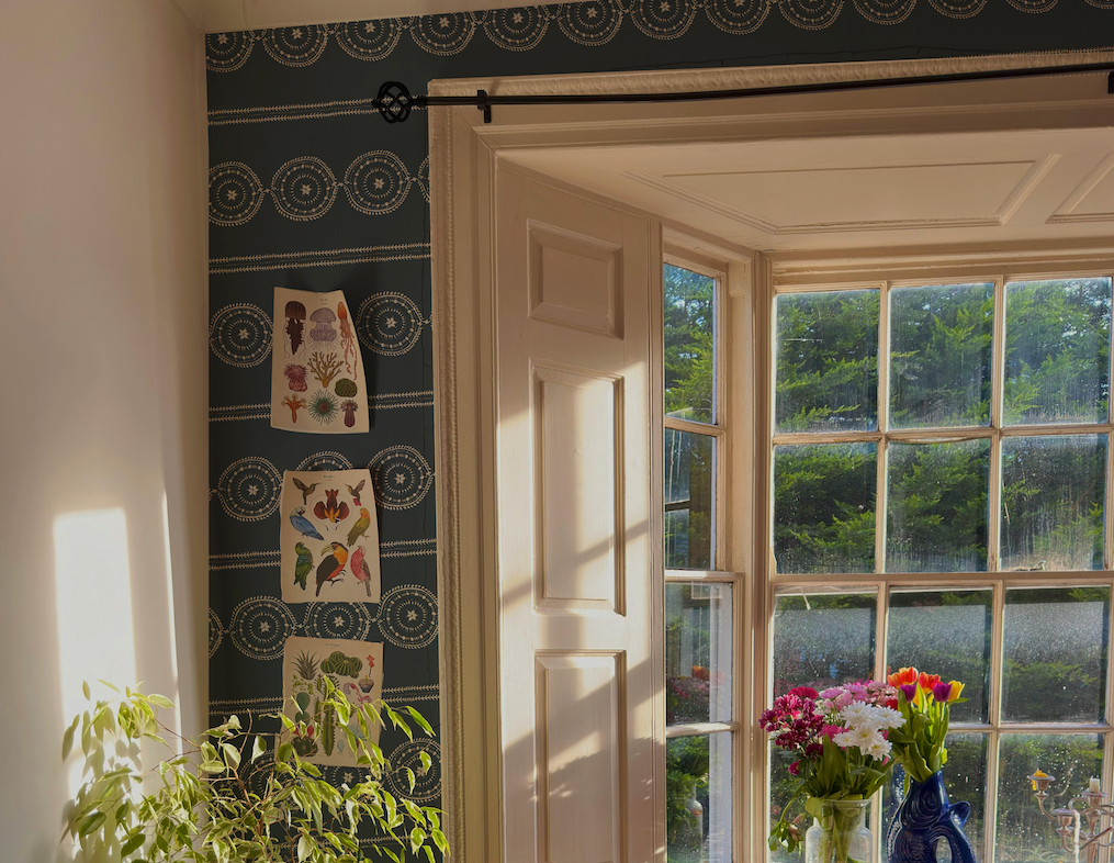 Sunlit room with a large window, decorative wall paper, and a vase of flowers.