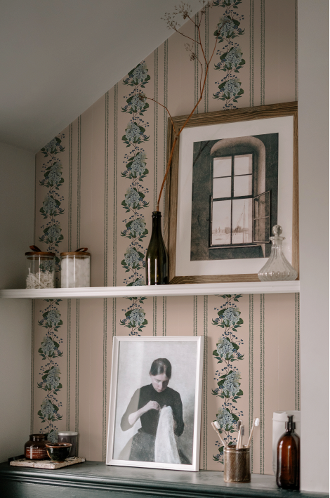 Decorative wall with framed pictures, candles, and a bottle against patterned wallpaper.