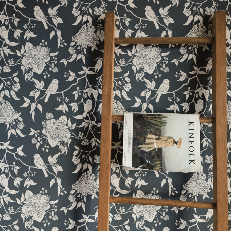 Wooden ladder with a book titled 'KINFOLK' against a floral-patterned wall.