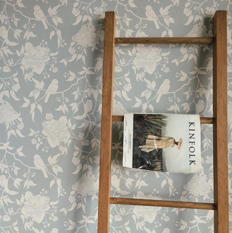Wooden ladder with a book titled 'KINfolk' against a floral wallpapered wall.