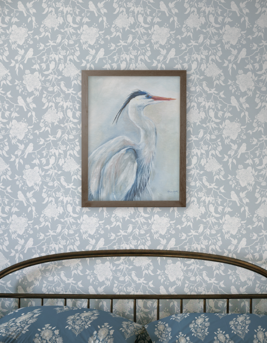 Framed painting of a heron on a floral wallpapered wall above a bed with matching bedding.