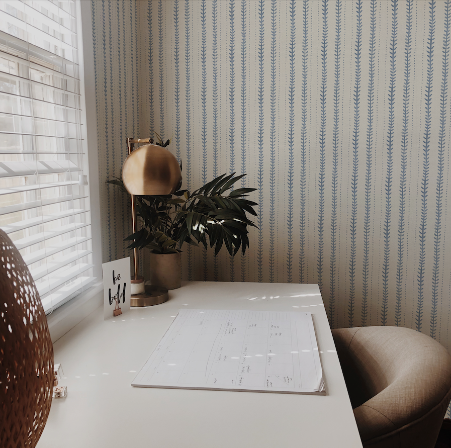 Home office with a desk, chair, plant, and calendar against a patterned wall.