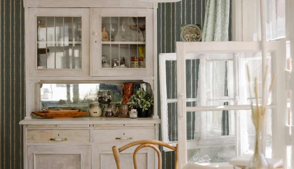 Kitchen interior with a white hutch, wooden chair, and window with curtains.
