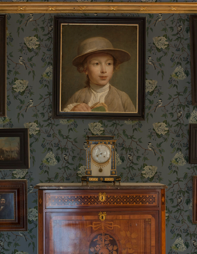Portrait of a young boy in a hat on a wall with decorative wallpaper and a wooden cabinet.