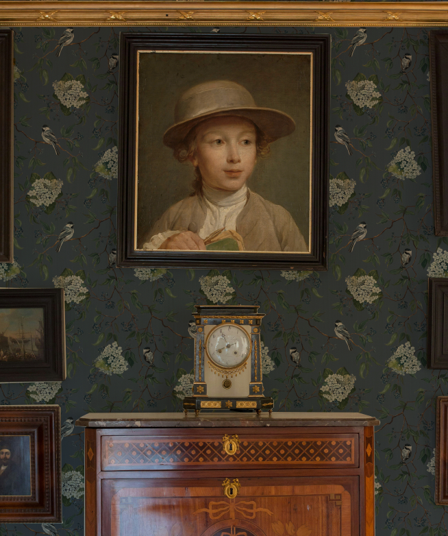 Portrait of a young boy in a hat on a wall with decorative wallpaper and a wooden cabinet.