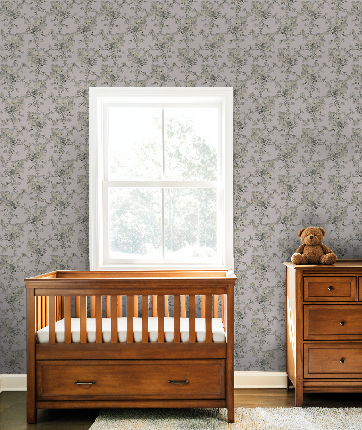 Wooden crib and dresser in a nursery with floral wallpaper and a window.