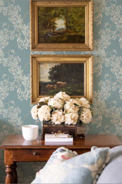 Two framed paintings above a wooden table with flowers and books against a patterned wall.