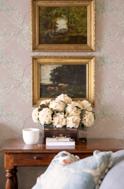 Two framed paintings above a wooden table with flowers and books, against a floral wallpapered wall.