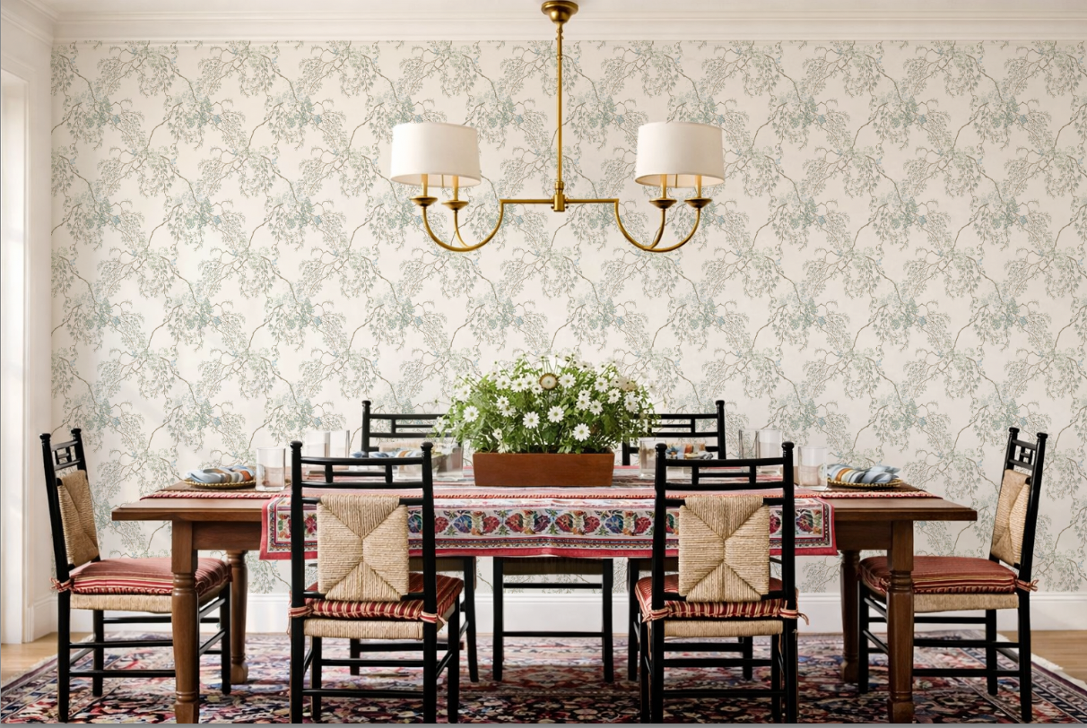A styled dining room featuring a dark wood dining table with six black chairs that have woven beige seat cushions, set on a patterned oriental rug against a wall with light green and white floral wallpaper. A brass double-arm chandelier with white drum shades hangs above.