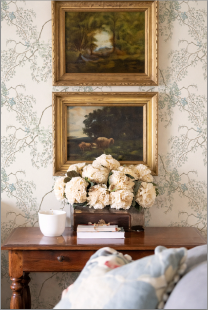Two framed paintings above a wooden table with flowers and books, against a patterned wall.