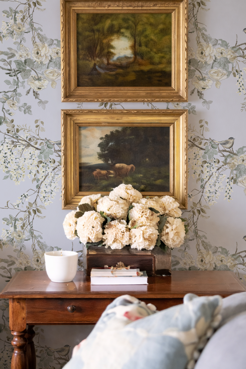 Decorative setting with floral arrangement, books, and paintings on a wall with floral wallpaper.