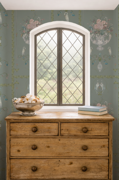 Wooden dresser with a bowl of seashells and books in front of a decorative window with patterned wallpaper.