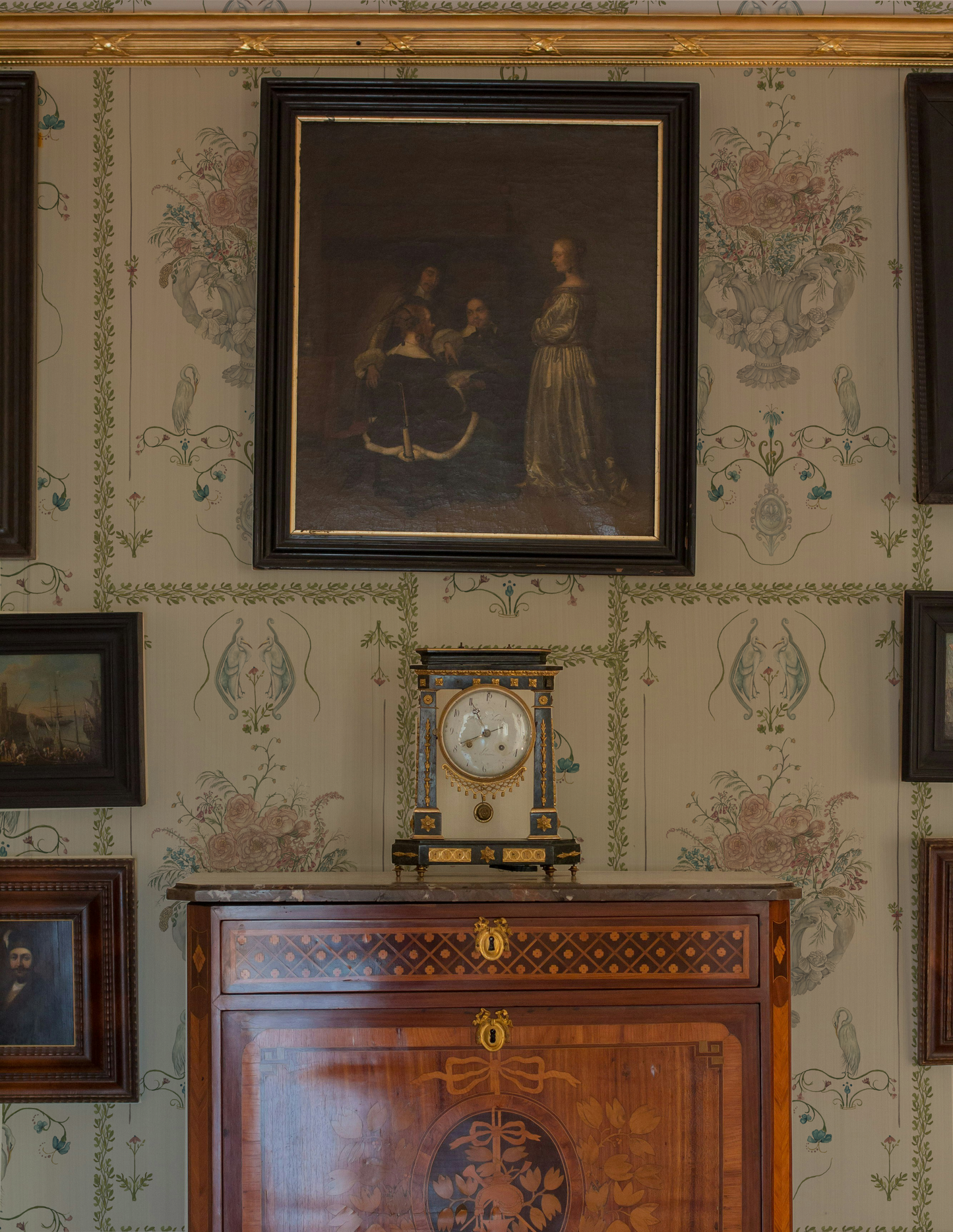 Decorative wall with framed portrait, clock, and wooden cabinet against ornate wallpaper.