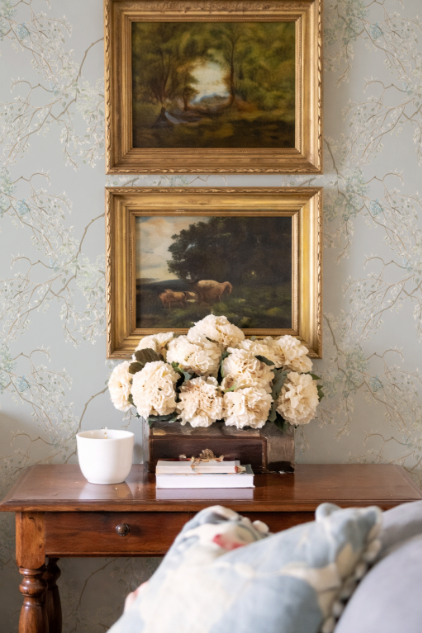 Two framed paintings above a wooden table with flowers and books, against a patterned wall.