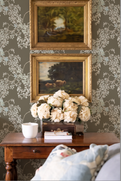 Two framed paintings above a wooden table with decorative items against a floral wallpapered wall.