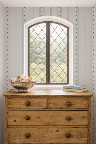 Wooden dresser with a bowl of seashells and books in front of a decorative window with striped wallpaper.